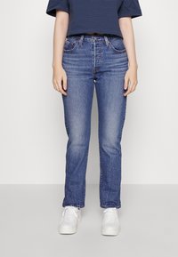 Jeans de mezclilla azul claro con corte recto, que presentan un desvanecimiento sutil y dos bolsillos frontales, combinados con una camiseta a rayas azul marino y blanca y zapatillas blancas.