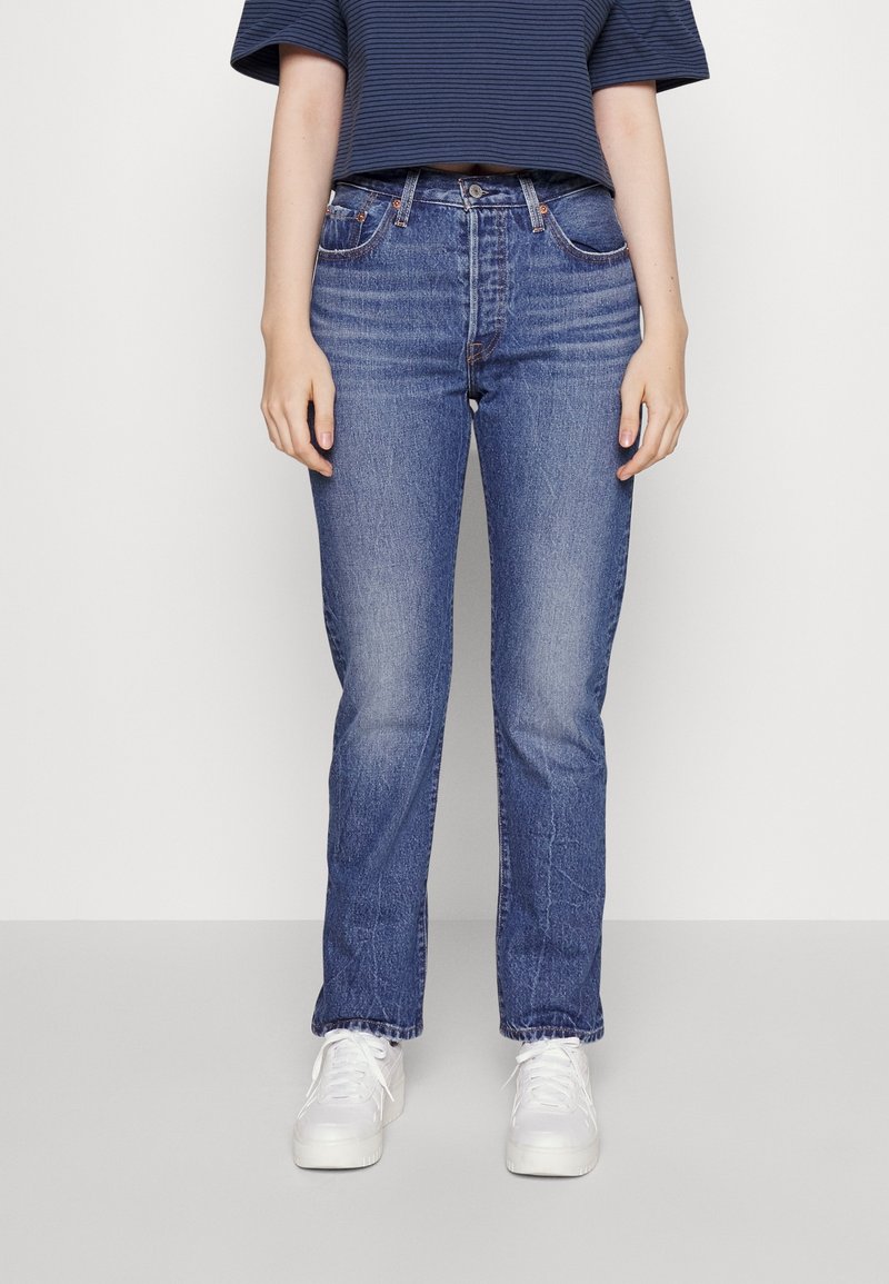 Jeans de mezclilla azul claro con corte recto, que presentan un desvanecimiento sutil y dos bolsillos frontales, combinados con una camiseta a rayas azul marino y blanca y zapatillas blancas.