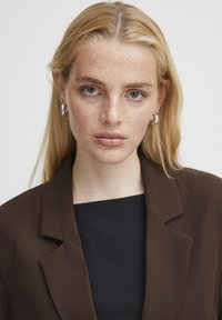 Blazer marron avec un col structuré sur un haut noir à côtes. Le mannequin a de longs cheveux blonds et des boucles d'oreilles en argent, sur un fond neutre.