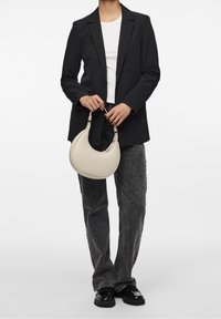 Jasno beżowa skórzana torebka o zakrzywionym kształcie i krótkich, mocnych uchwytach, trzymana przez osobę w czarnym żakiecie i szarych dżinsach.