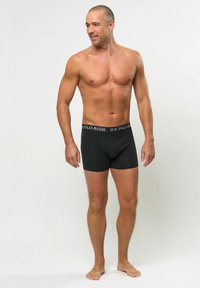 Svarta bomullsboxershorts med midjeband som har en U.S. Polo Assn.-logotyp. Modellen står barfota och visar upp en figurnära design och en enkel stil.