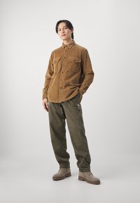 Brown corduroy shirt with two chest pockets, paired with green cargo pants and light brown boots. Neutral background provides contrast.