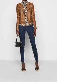 Bruine leren motorjack over een bloemenmocknecktop, gecombineerd met donkere skinny jeans en zwarte sandaaltjes met hakken, terwijl ze een kleine zwarte tas vasthoudt.