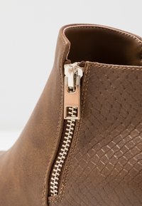 Bottine à cheville en similicuir marron avec un côté texturé, une fermeture éclair en métal argenté et un bord supérieur lisse. Accentuée d'un motif tissé.