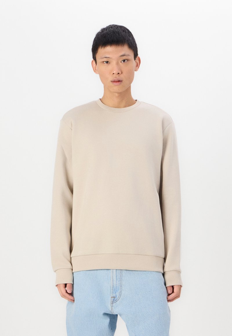 Beige sweatshirts i ett mjukt material med rund hals, långa ärmar och ribbade ärmar, parat med ljusblå jeans.