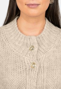 Cardigan a maglia beige con colletto a coste, presenta due bottoni dorati nella parte anteriore. Il tessuto appare morbido e con una texture piacevole.