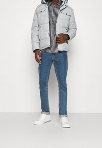 Giacca imbottita grigia chiara con cappuccio, bottoni neri, camicia a quadri bianca e blu navy, jeans blu e sneakers bianche.