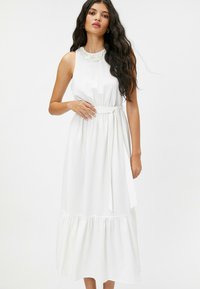 Robe midi blanche en tissu texturé, col montant, avec des broderies au niveau du col et une ceinture à nouer à la taille ; ourlet à volants.