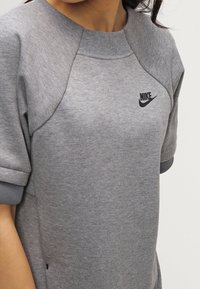 Grå sweatshirt med rund halsringning, korta ärmar och en svart Nike-logotyp på bröstet. Tyget verkar mjukt med en slät yta.