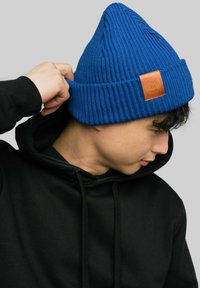 Niebieska dzianinowa czapka z wierzchołkiem, o żebrowanej fakturze, z brązową skórzaną łatą. Noszona na czarnej bluzie z kapturem.