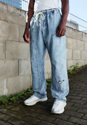 Homme portant un pantalon en jean bleu clair à jambes larges avec logo Nike et des baskets blanches, debout sur le trottoir près d'un mur en béton.