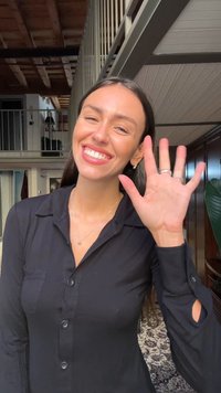 Mulher vestindo uma camisa preta de botão com as mangas arregaçadas, sorrindo e acenando. O fundo apresenta um teto de madeira e plantas de interior.