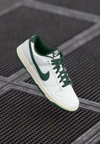 Biela a zelená teniska Nike s šnúrkami, plávajúca nad kovovou mriežkovou plochou.