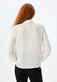 Blusa bianca a maniche lunghe con colletto, realizzata in un tessuto leggero e morbido; la vista sul retro mostra un orlo dritto e una leggera drappeggiatura sulle maniche.