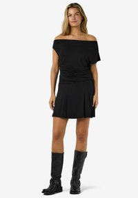 Kobieta w czarnej bluzce z odkrytymi ramionami, czarnej plisowanej spódnicy i czarnych skórzanych kozakach do kolan, stojąca na jednolitym białym tle.