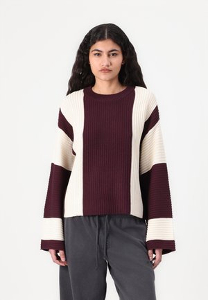 Maroon and cream striped sweater with ribbed texture, long flared sleeves, and a round neckline, styled with casual dark pants.