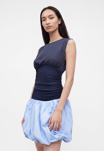 Femme aux longs cheveux foncés portant une robe sans manches avec un haut ajusté sombre et une jupe bouffante bleu clair, debout devant un fond blanc uni.