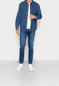 Camisa de mezclilla en azul oscuro, con un diseño de botón y costuras en contraste. Combinada con jeans azules y zapatillas blancas.