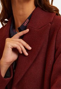 Femme ajustant le col d'un manteau en laine rouge foncé, portant une chemise à motifs bleu foncé en dessous avec du texte sur le col.