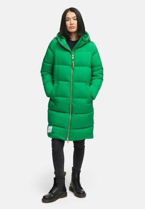 Femme se tenant debout avec les mains dans les poches, portant un manteau long en doudoune vert, un jean noir et des bottes noires à lacets sur un fond blanc.
