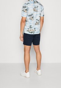 Chemise à manches courtes pour homme en bleu clair avec un imprimé de palmier et d'île, accompagnée de shorts navy et de baskets blanches.