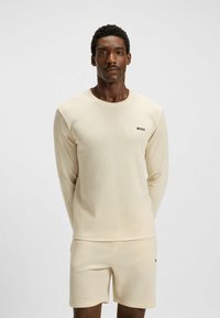 Beige set bestaande uit een lange mouwen top en shorts, gemaakt van textuurmateriaal. Bevat een klein zwart 'BOSS' logo op de borst. Eenvoudig, getailleerd ontwerp.