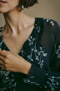 Blusa floreale scura con maniche lunghe, scollatura arricciata e polsini elastici. Presenta delicati motivi di fiori bianchi e rosa su uno sfondo blu navy.