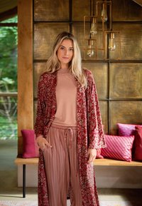 Jonge vrouw met lang blond haar, gekleed in een rode bloemenrobe over een roze top en gestreepte broek, staand binnen bij een houten bank met rode kussens.