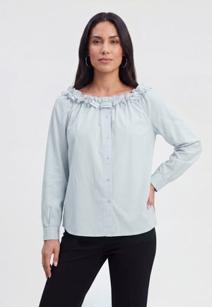 Frau mit langen dunklen Haaren, die eine hellblaue geraffte Schulterfreie Bluse und schwarze Hose trägt, steht mit einer Hand auf der Hüfte vor weißem Hintergrund.