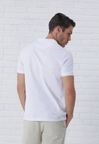 White cotton polo shirt with a classic collar, short sleeves, and straight hem, worn by a man seen from the back against a brick wall.