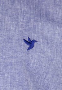 Tela azul con una textura sutil; presenta un logotipo de colibrí bordado en azul oscuro ubicado a la izquierda.