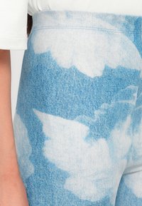 Pantalones de denim de tiro alto con un lavado azul claro y patrones de nubes blancas esponjosas. El material es suave y presenta detalles de costura visibles.