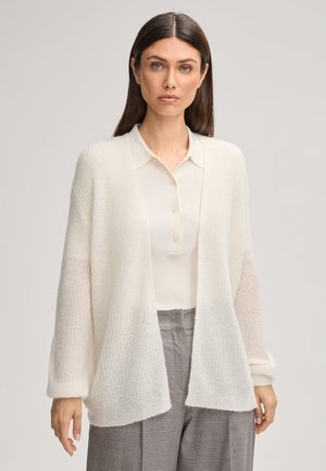 Donna che indossa un cardigan bianco a maglia sopra una camicia bianca con colletto e pantaloni grigi a quadri, in piedi contro uno sfondo chiaro e uniforme.