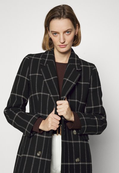 Cappotto a quadri neri con motivo a check grigio, fronte doppiopetto, colletto a punte e dettagli con bottoni, indossato sopra un maglione di maglia marrone.