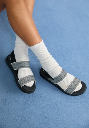 Fekete szandál csíkos anyagú pántokkal, fehér bordázott zoknikkal viselve. Kék háttér előtt, kiemelve egy laza megjelenést.