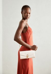 White textured handbag with a gold clasp, held by a person in a rust-colored sleeveless dress. The bag features a chain strap and geometric pattern.