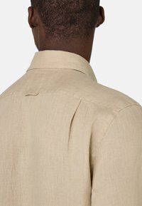 Camicia in lino beige chiaro con colletto classico, pieghe sul retro e un delicato motivo testurizzato. Messa a fuoco sulla semplicità e su un tessuto traspirante.