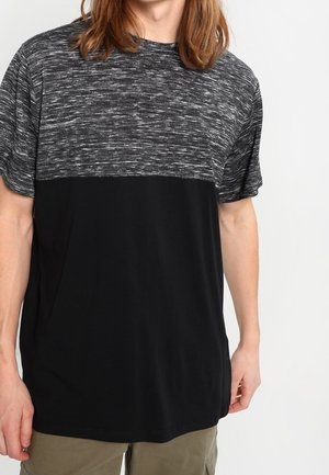 Mann trägt ein kurzärmeliges T-Shirt mit grauem und schwarzem Farbblock und olivgrüner Hose, steht vor einem einfarbigen weißen Hintergrund.