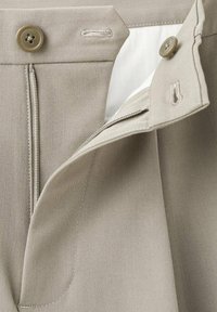 Nærbillede af beige bukser med knappet talje, synlig lynlås og stofklap, der er delvist åben.