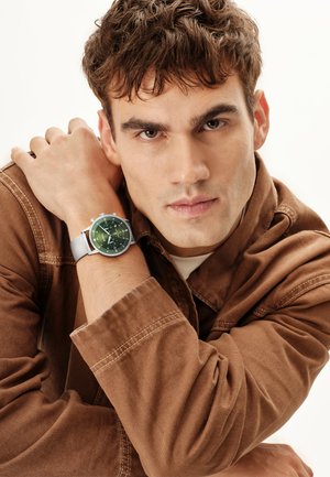 Jeune homme en veste marron reposant son menton sur ses bras croisés, portant une montre en maille argentée avec un cadran vert à son poignet gauche.