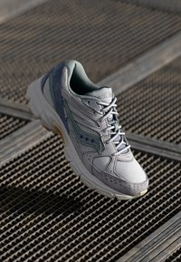 Chaussure de sport grise avec des panneaux en maille et en daim, accents verts, bout arrondi et semelle texturée. Elle possède des lacets et un logo sur le côté.