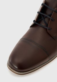 Zapato de cuero marrón con punta redondeada, costuras decorativas y cordones oscuros. Textura suave con un brillo sutil y una suela de goma ligera.