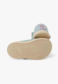Zapatilla de lona azul claro con suela de goma beige. Presenta un parche de logo circular en la parte trasera y rayas blancas texturizadas en la suela.