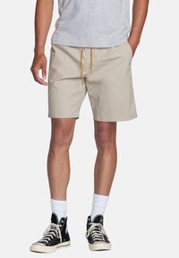Shorts beiges avec un cordon de serrage jaune, présentant une texture lisse et des poches latérales, associés à des baskets montantes noires.