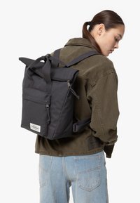 Black nylon roll-top backpack with front pocket, zipper closures, and adjustable straps. Features logo patch on the front.