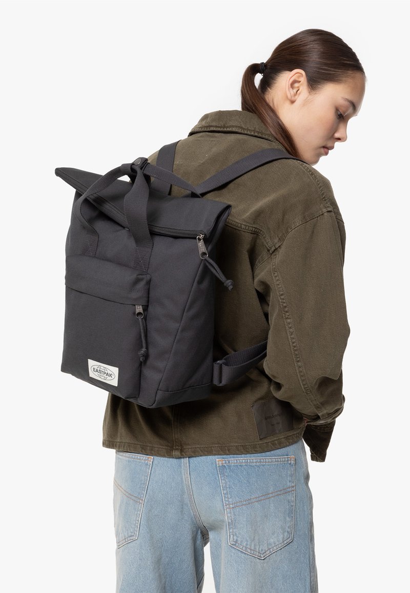 Black nylon roll-top backpack with front pocket, zipper closures, and adjustable straps. Features logo patch on the front.