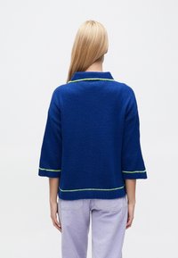 Nadmiernie duży niebieski dziany sweter z kołnierzem, rękawami do łokcia oraz kontrastującymi zielonymi szwami na krawędzi i mankietach rękawów.