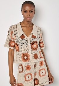 Häkelcardigan in Beige mit orangefarbenen und braunen kreisförmigen Mustern. Kurze Ärmel, knöpfbare Vorderseite und Kragen. Strukturiertes und offenes Gewebedesign.