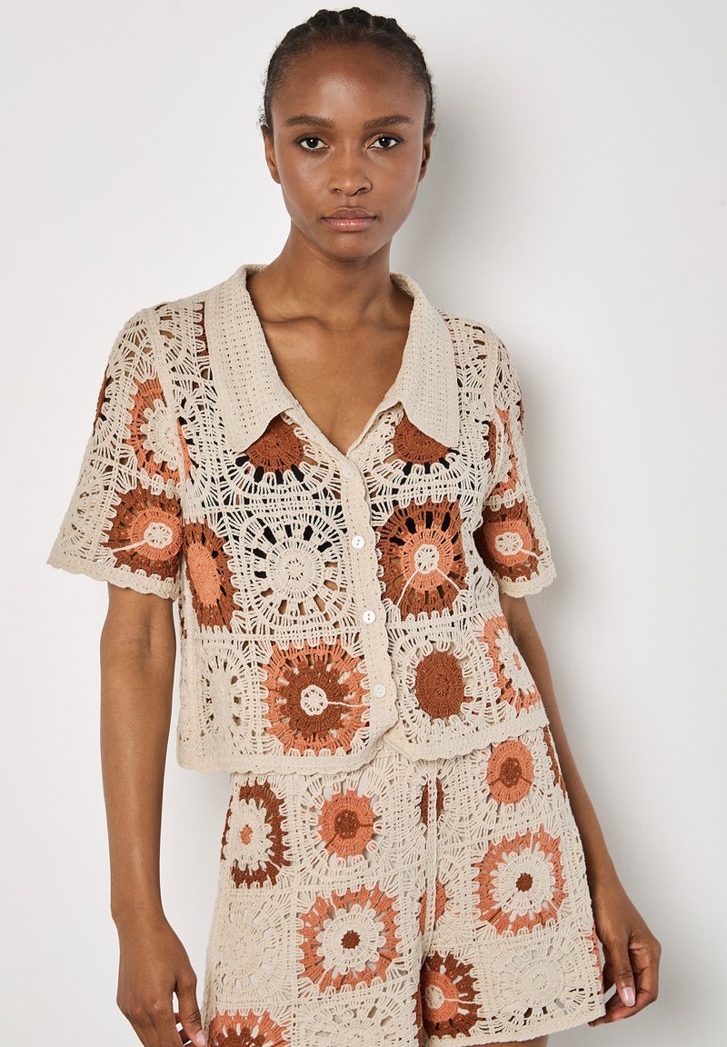 Häkelcardigan in Beige mit orangefarbenen und braunen kreisförmigen Mustern. Kurze Ärmel, knöpfbare Vorderseite und Kragen. Strukturiertes und offenes Gewebedesign.
