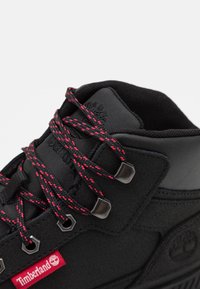 Czarne buty trekkingowe z teksturowaną syntetyczną cholewką, czerwono-czarnymi sznurowadłami w wzór, metalowymi oczkami i czerwoną etykietą Timberland.